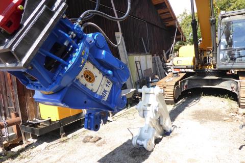 Trevi MK15 KIT FR Wechselbacke – auf dem Boden liegend, im Hintergrund das MK-15-Grundgerät am Baggerausleger, Werkstatthof mit Holzscheune